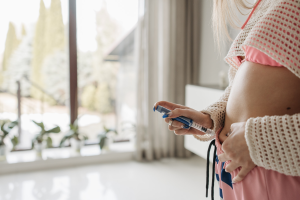 Person injecting medication into abdomen at home, wearing casual clothing, next to large window with plants