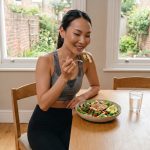 Person enjoying a healthy salad at home with a glass of water on a wooden table in a bright dining area